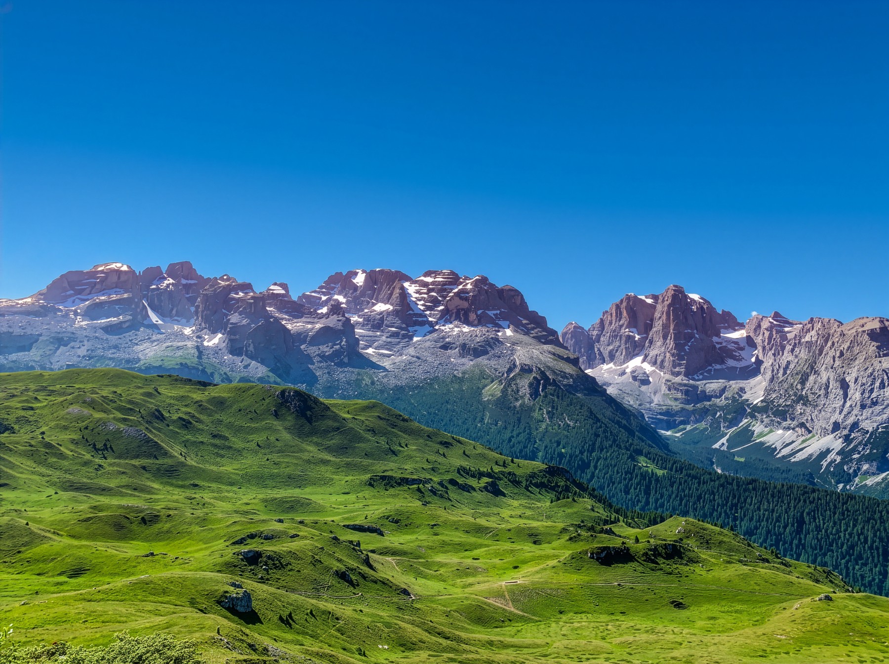 Jagged snow-dusted limestone peaks of the Brenta Dolomites rising above green alpine meadows under a deep blue sky