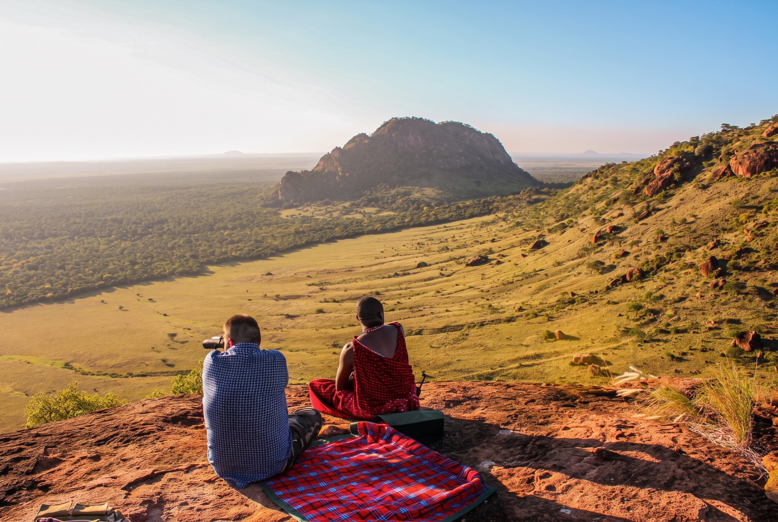 Luigi sits beside Pashiet, a Maasai friend, on a rocky ridge at golden hour, looking through binoculars over a vast green valley and volcanic hills below